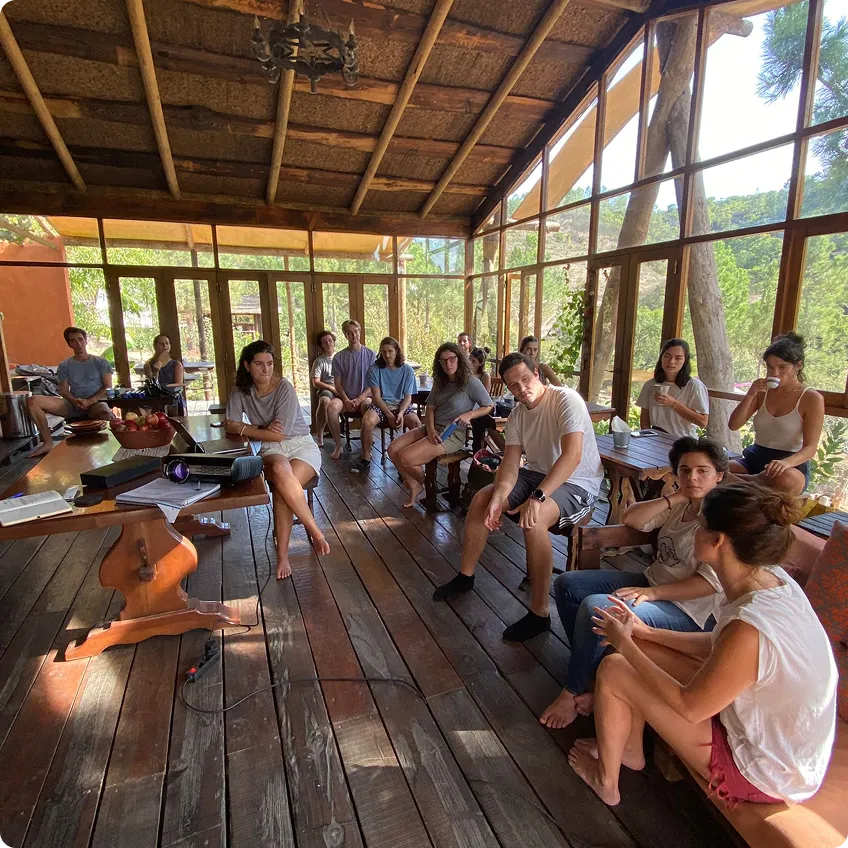 Group of young adults seated on wooden floor and benches in a sunlit glass-walled room with trees outside.