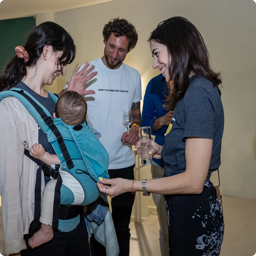 Three adults smiling and interacting with a baby in a teal carrier at an indoor gathering.