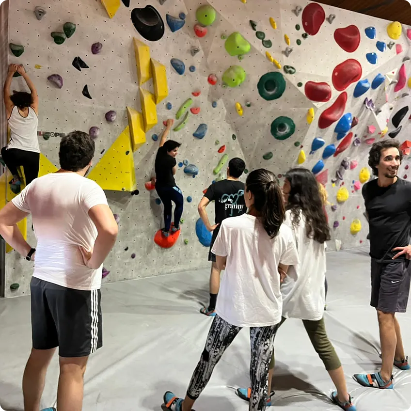 Group of people indoor bouldering on colorful climbing holds on a grey wall with padded floor.