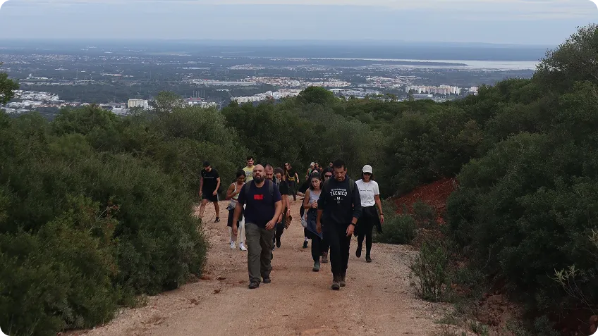 Group of hikers walking up a dirt trail surrounded by green bushes with a city and water view behind.