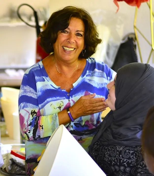 Smiling woman in a colourful blue top warmly touching the chin of a seated woman wearing a grey hijab.