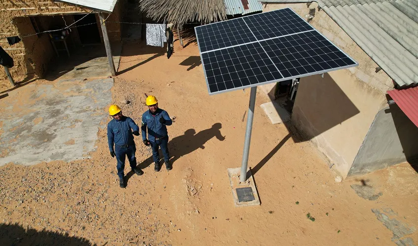 Two workers in blue uniforms and yellow helmets pointing near a mounted solar panel in a sandy yard.