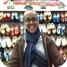 Smiling person wearing glasses, scarf, and jacket standing in front of a wall display of colorful shoes.