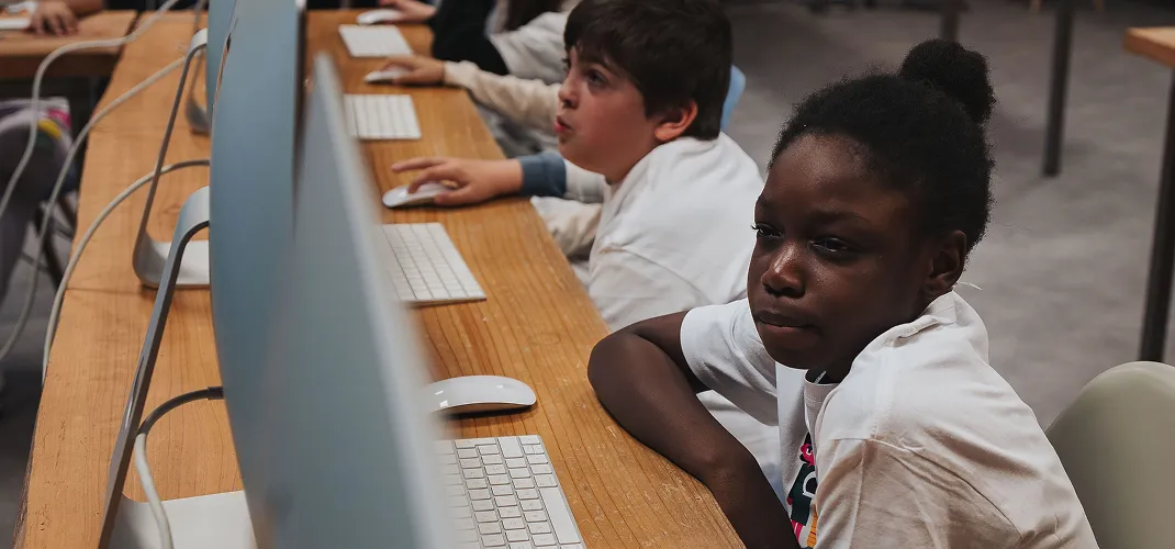 Children sitting at a wooden desk using desktop computers with keyboards and mice in a classroom.
