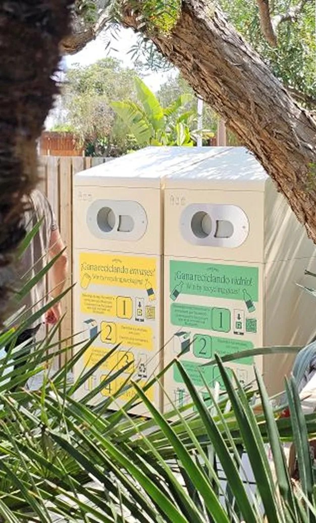 Two outdoor recycling bins labelled for glass and mixed recycling, surrounded by greenery.