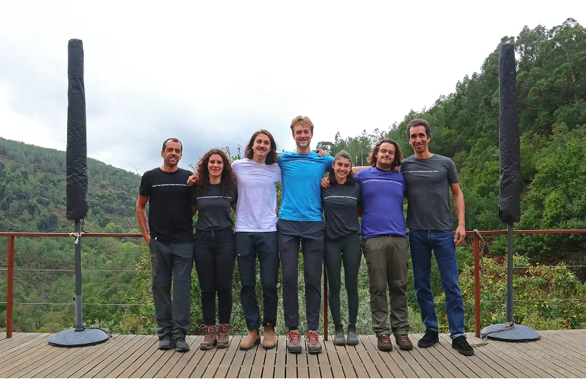 Seven people standing arm in arm on a deck with a forested hillside in the background.