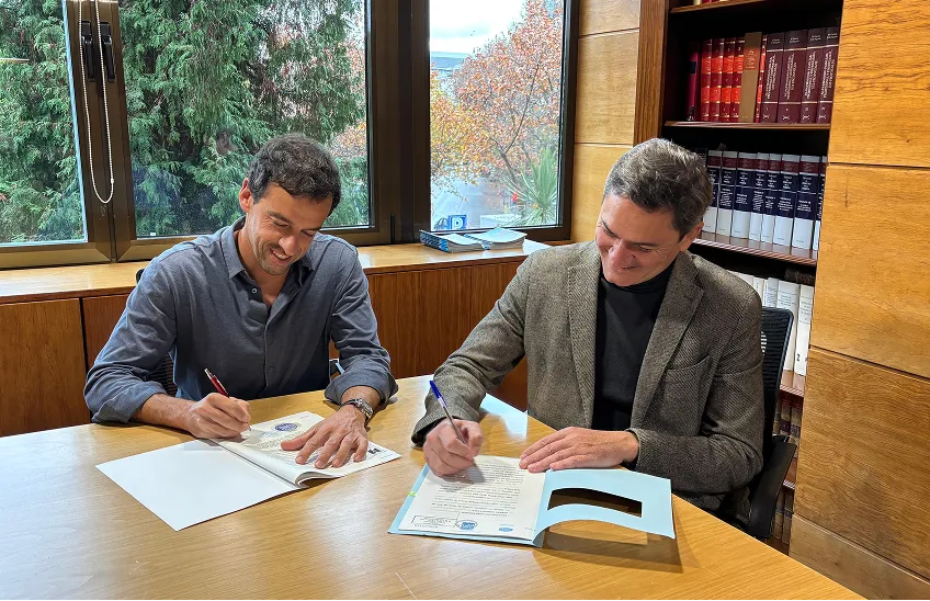 Two men seated at a wooden table signing documents in an office with bookshelves and large windows.