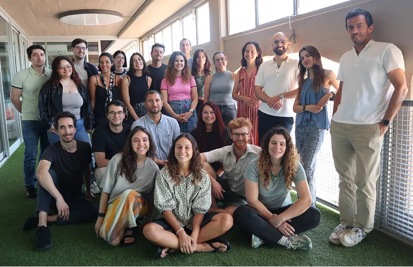 Group photo of 22 young adults smiling indoors on artificial grass under ceiling lights.