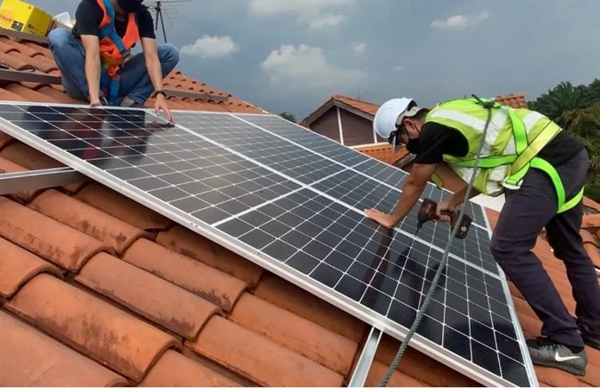 Two workers install solar panels on a tiled roof, wearing safety gear and harnesses.