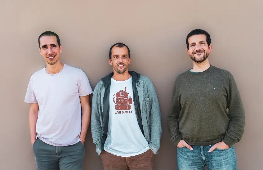 Three men standing side by side smiling against a plain light brown wall.