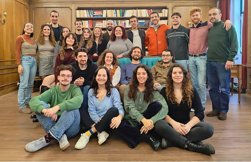 Group of 23 young adults smiling in a room with wooden walls and bookshelves.
