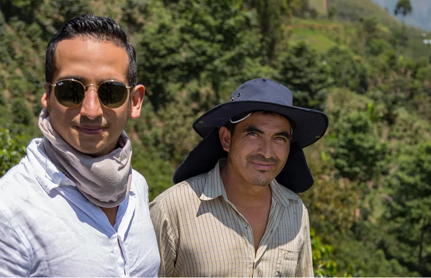 Two men outdoors in daytime; one wears sunglasses and scarf, the other a broad hat and plaid shirt.
