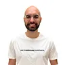 Bald man with glasses, beard, smiling wearing a white T-shirt with small text.