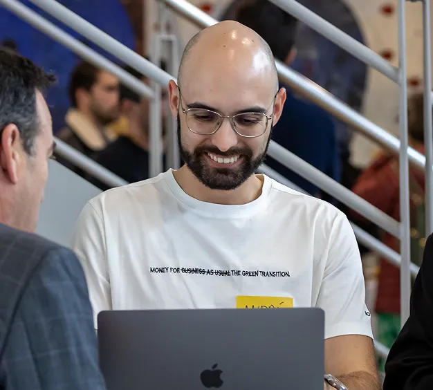 Smiling man with glasses and a beard working on a laptop in a busy indoor setting.