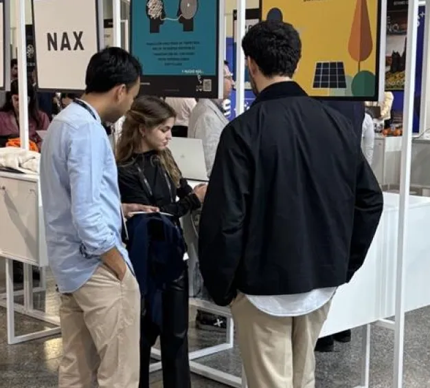 Three people standing and discussing inside a booth at an event with display boards overhead.