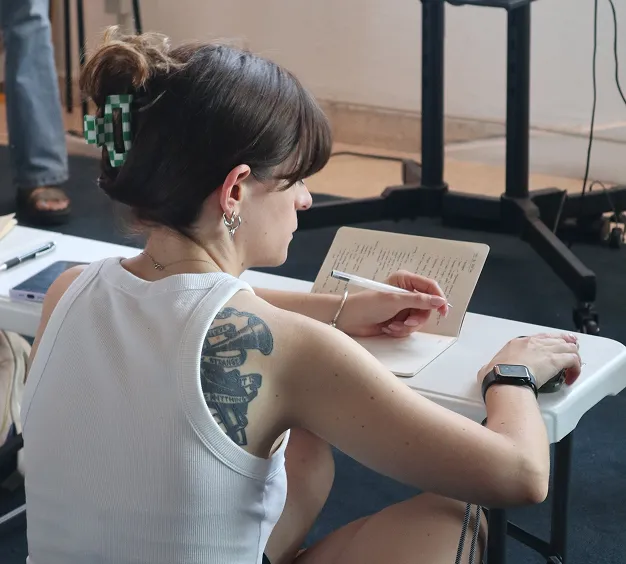 Woman with tattoo and green checkered hair clip writing notes in a notebook at a white table.