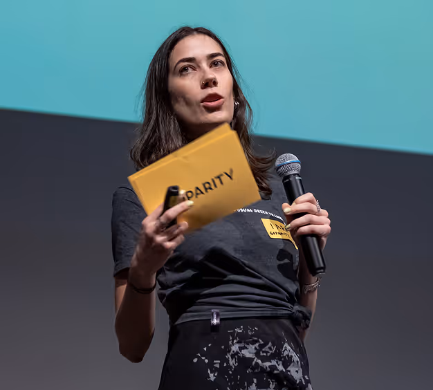 Woman holding a microphone and yellow card saying "PARITY" while speaking on stage.