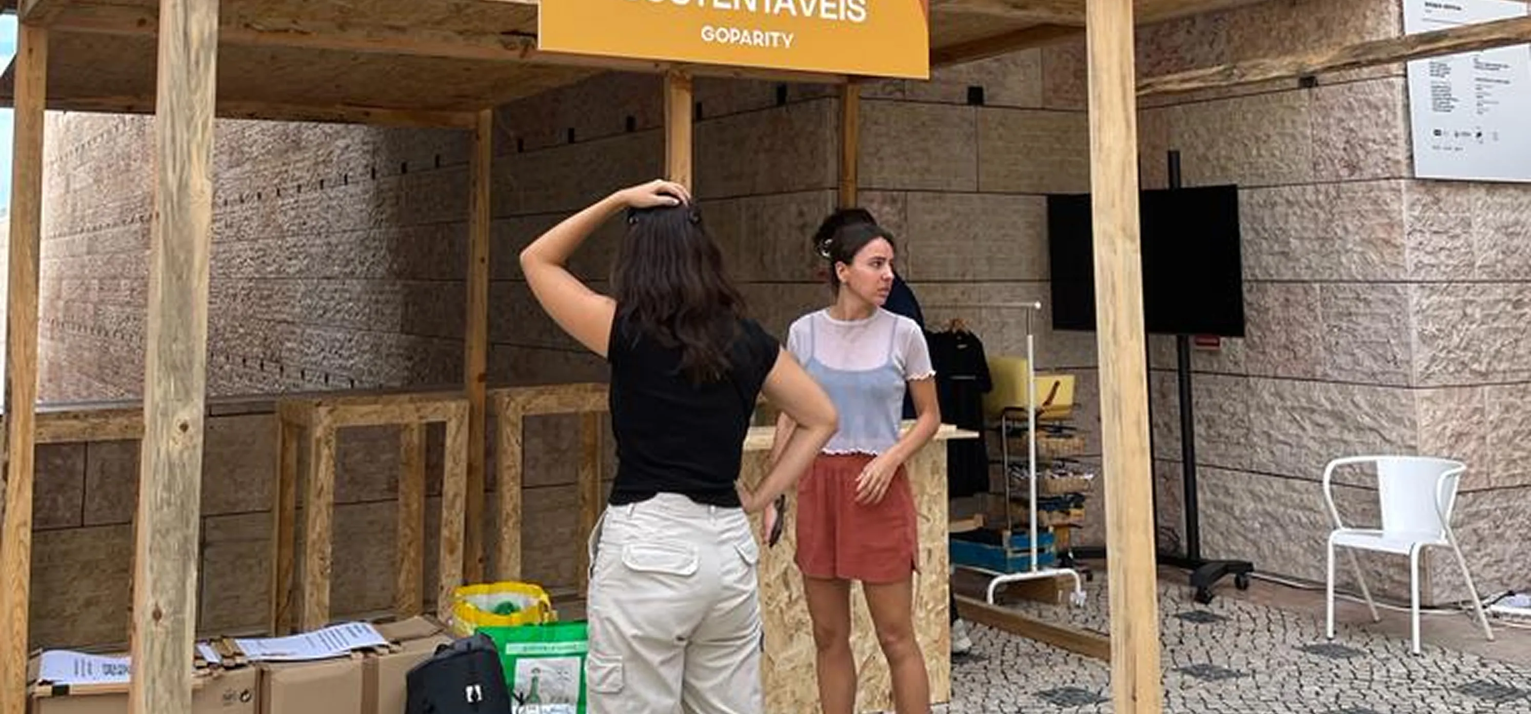Two women talking near a wooden booth with boxes and supplies on a cobblestone floor.