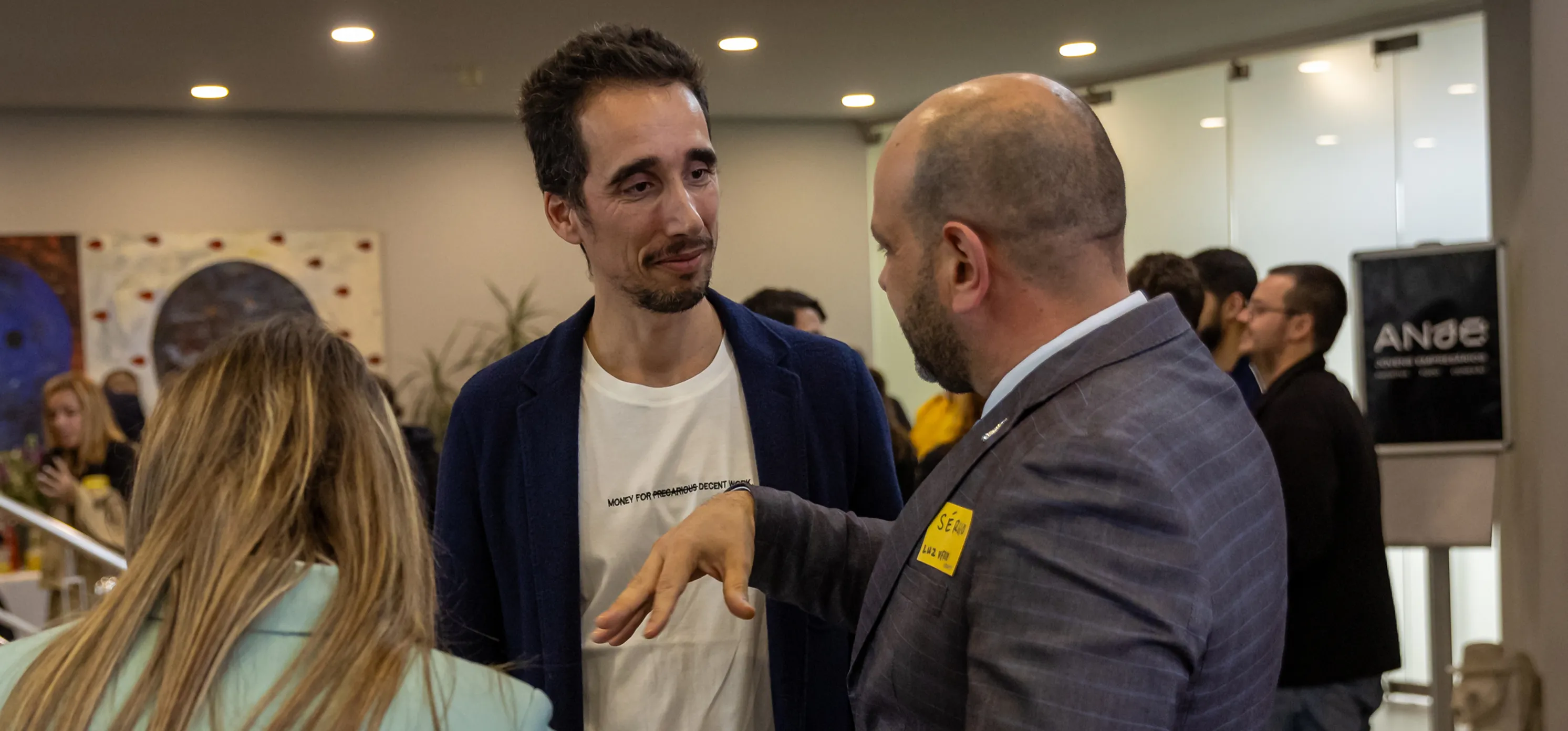Two men in conversation at an indoor event with other attendees blurred in the background.