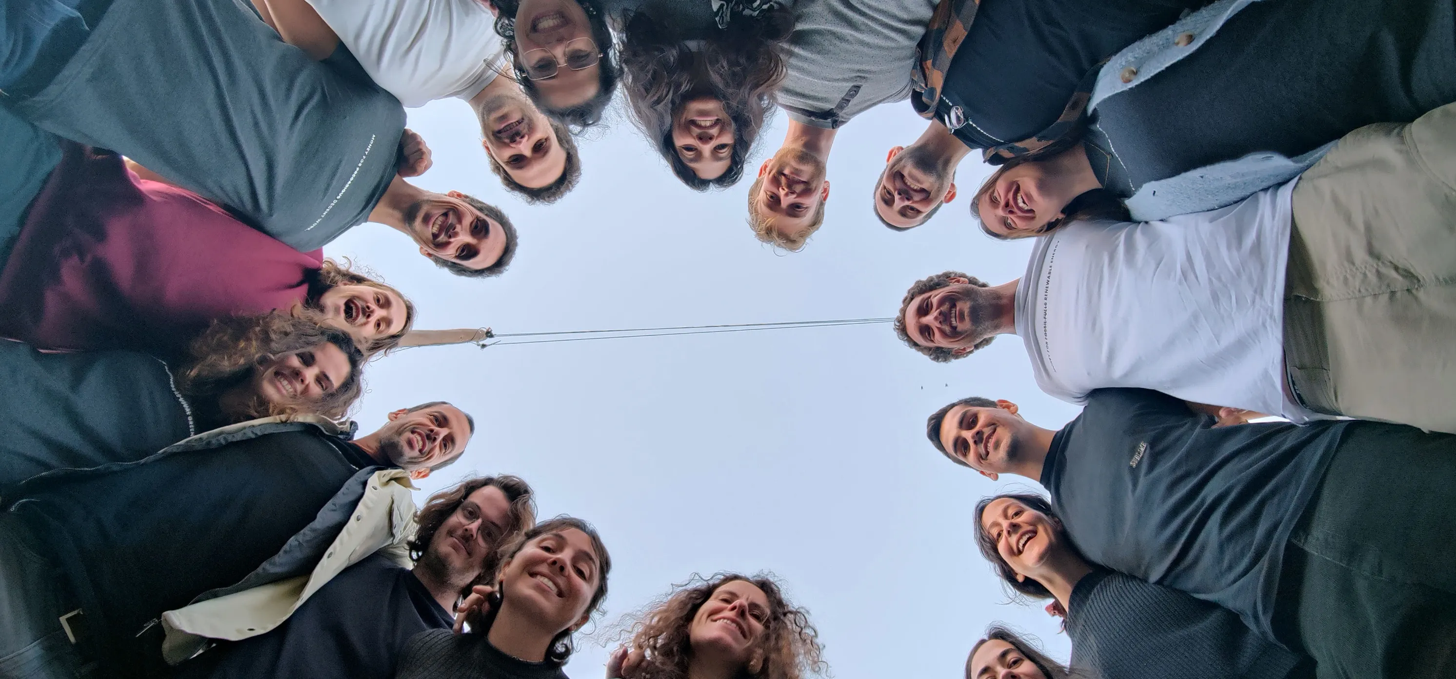 Group of people standing in a circle looking down at the camera against a clear sky.