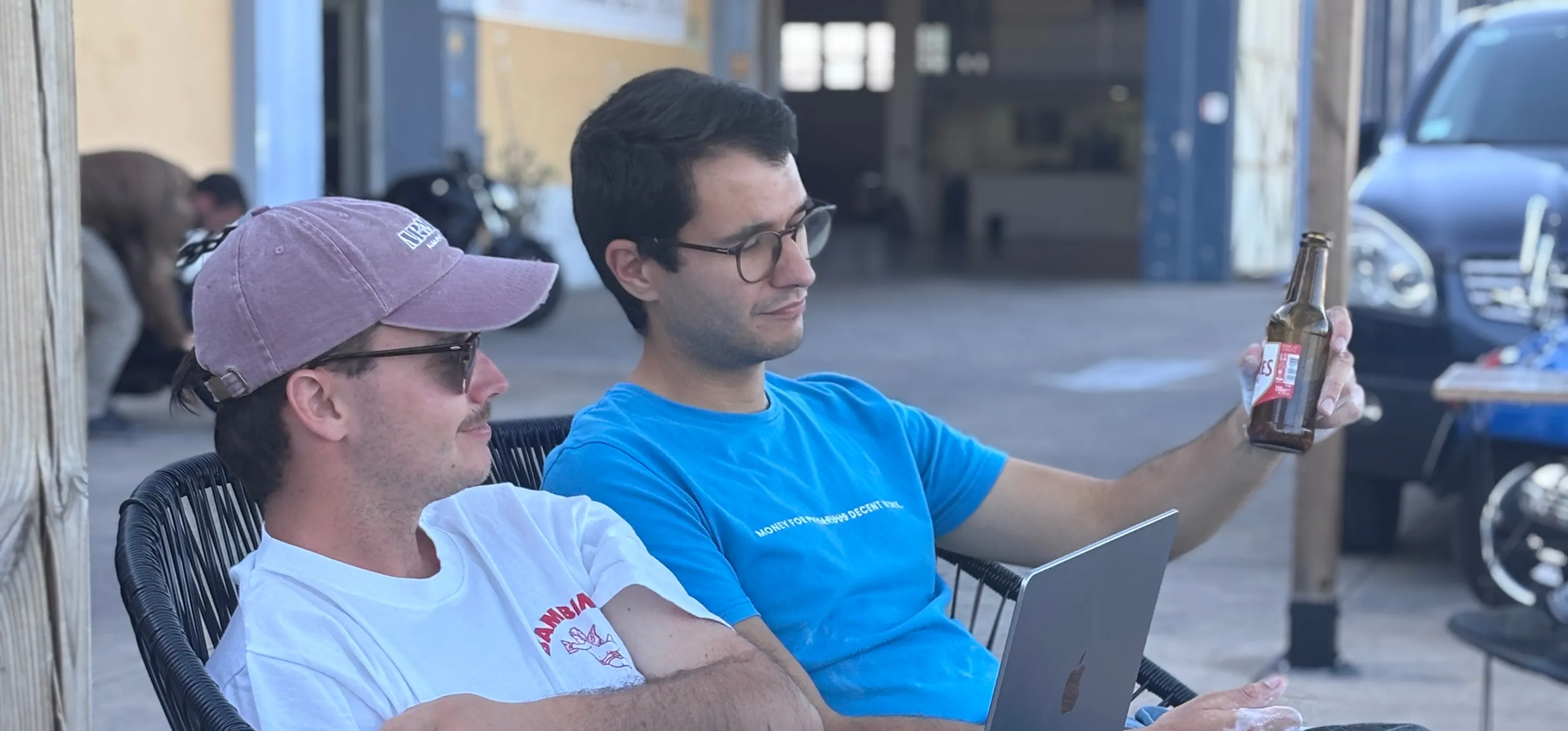 Two men sitting outdoors, one holding a beer bottle and the other wearing a cap and sunglasses.