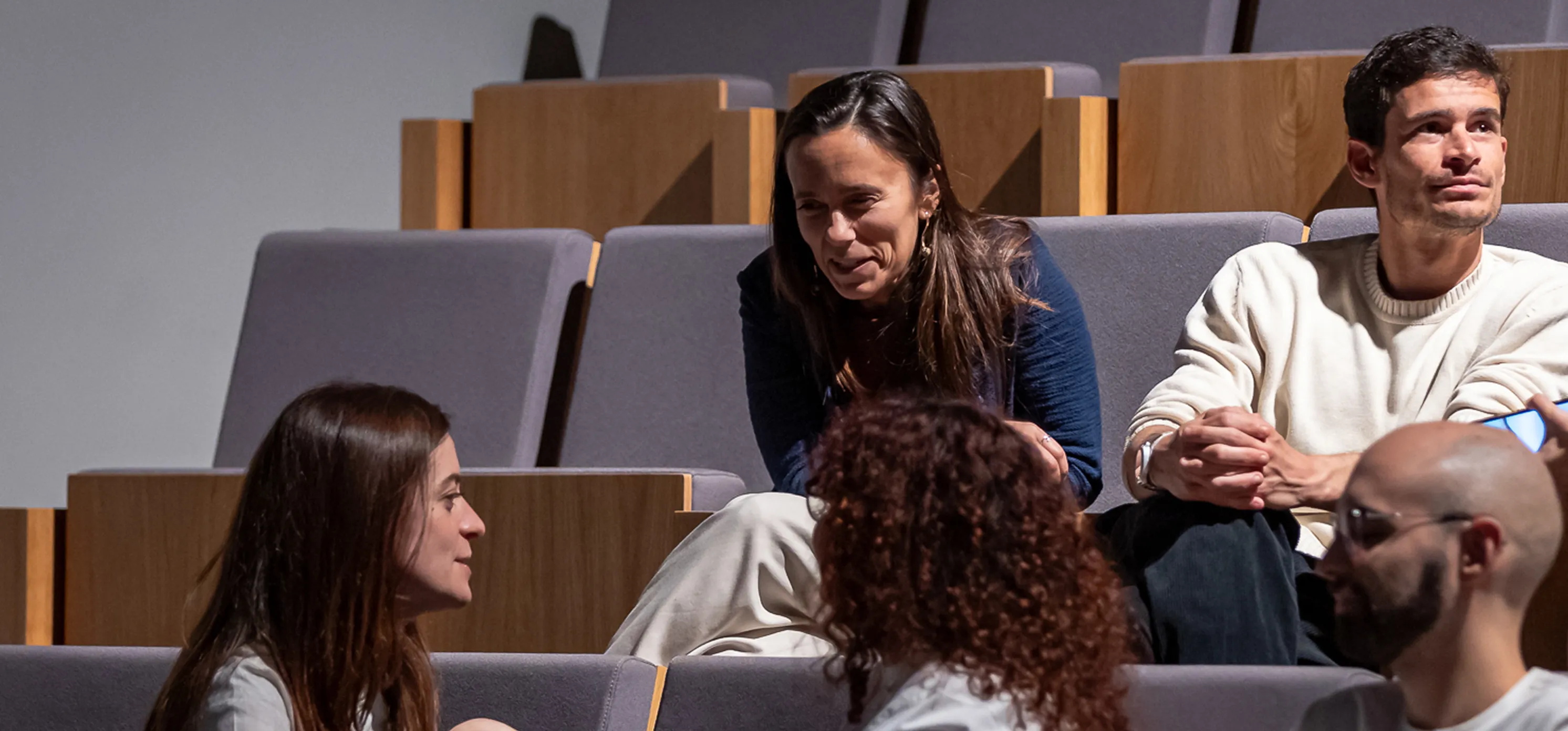 Four people sitting and talking in a tiered auditorium with wooden and grey seating.