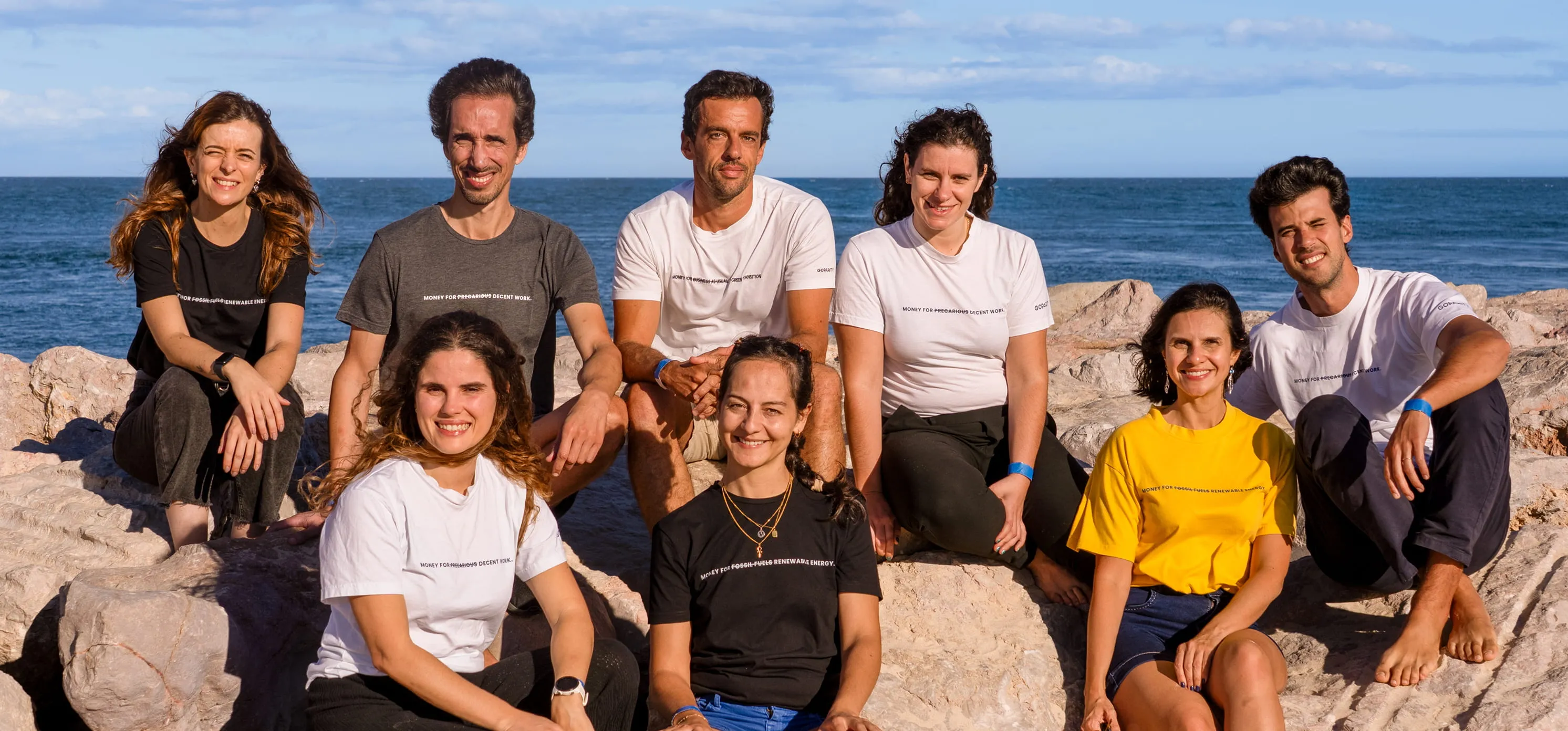 Group of eight adults sitting on rocks by the sea, wearing casual clothes and smiling at the camera.
