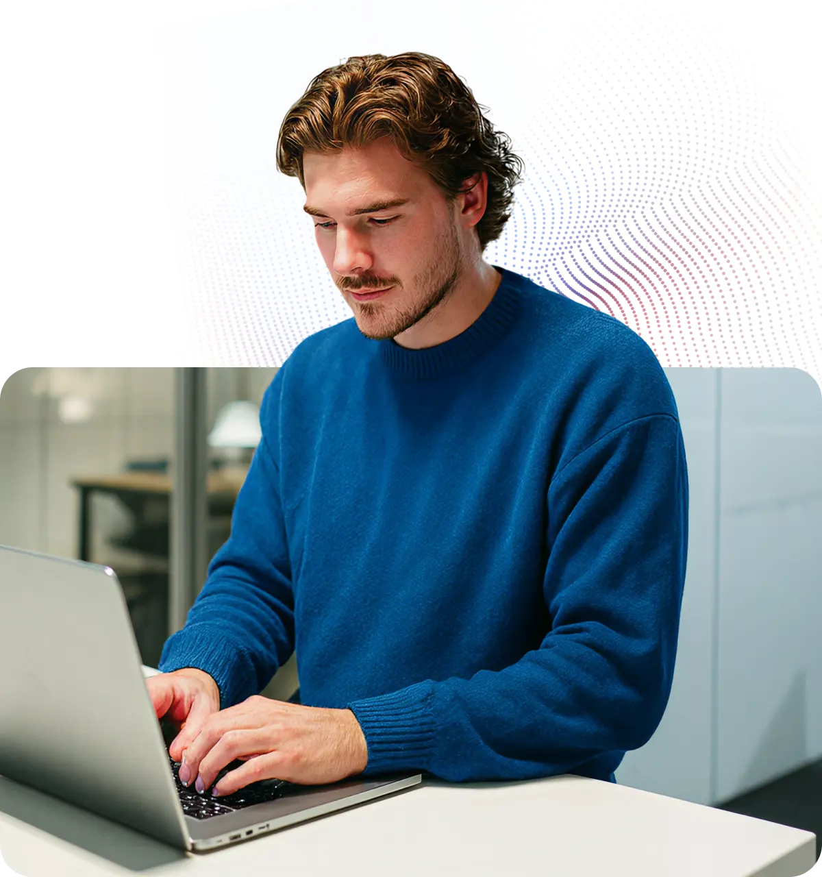Young man in a blue sweater typing on a laptop at a desk in an office setting.