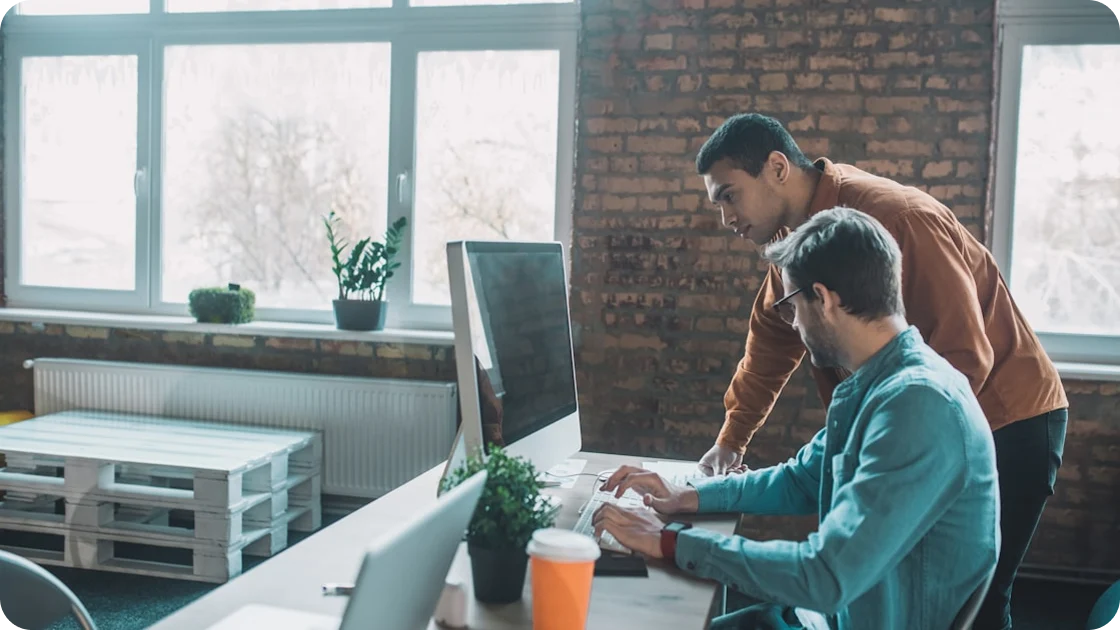Two men working together at a desk with a computer in a modern office with large windows and brick walls.