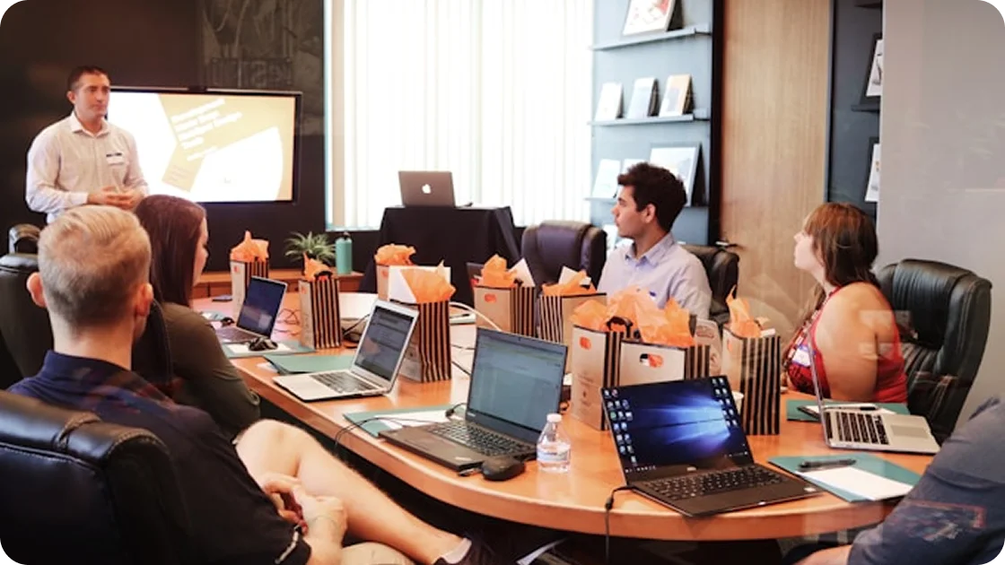 People sitting around a conference table with laptops listening to a man giving a presentation in a meeting room.