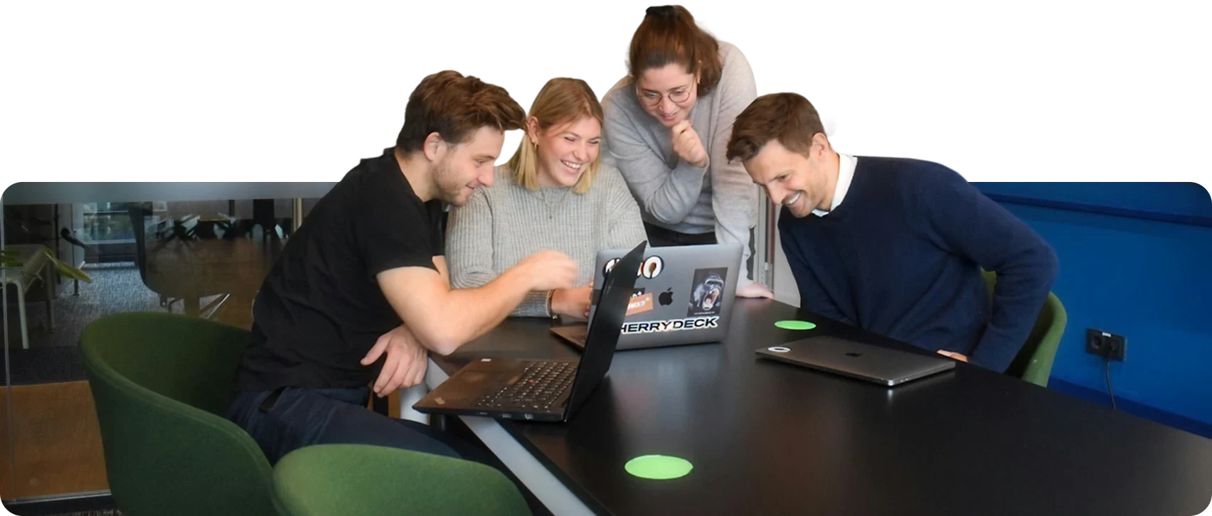 Four young professionals smiling and looking at two laptops while sitting around a table in an office.