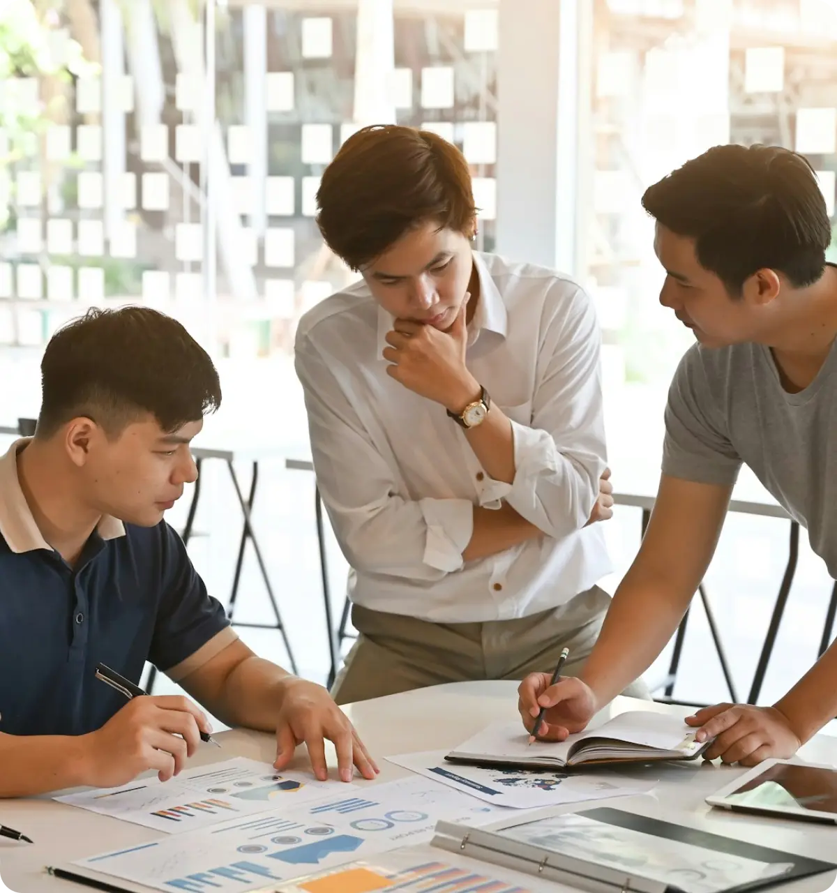 Three men collaborate over charts and notebooks at a table in a bright office.