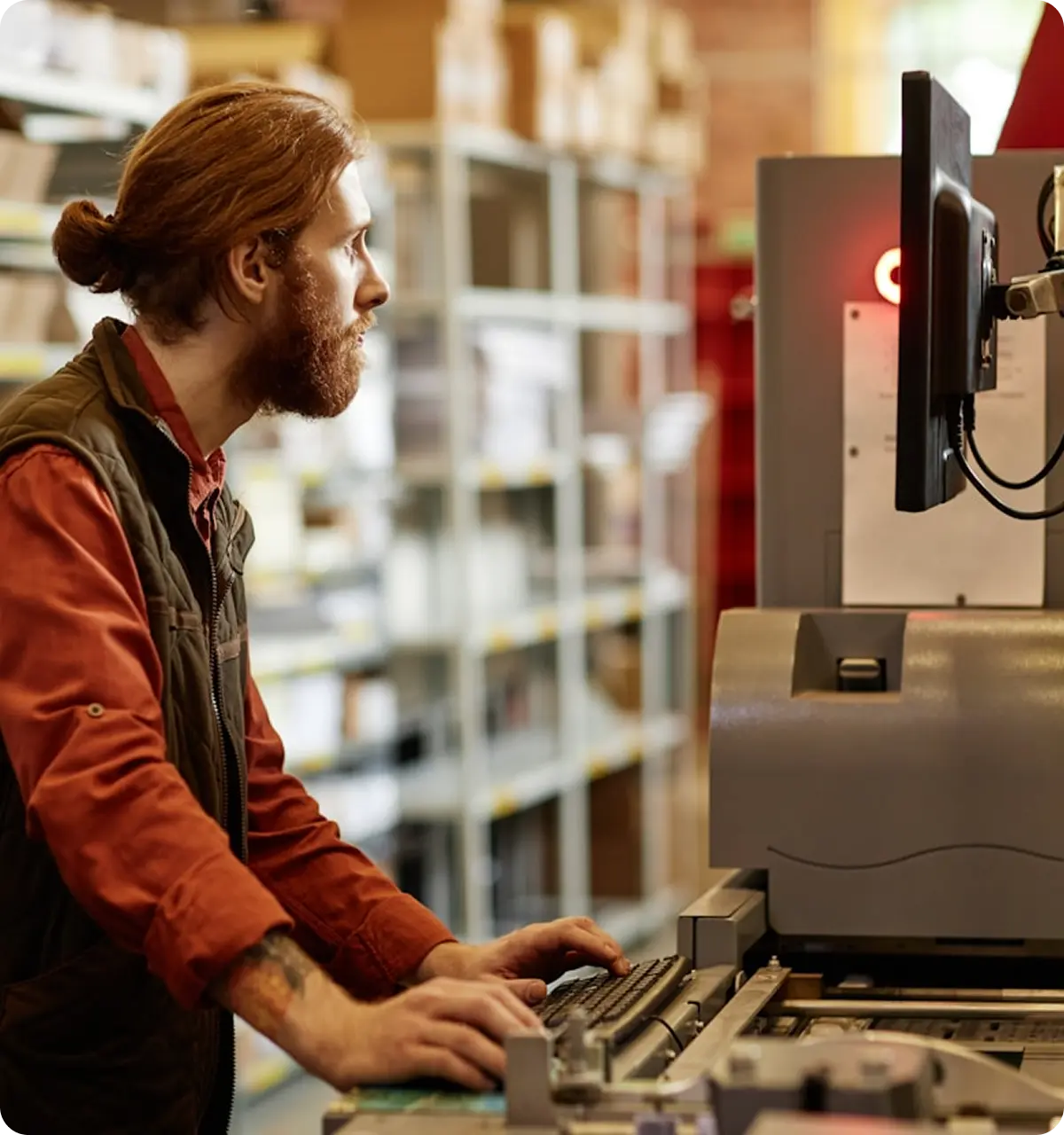 Man with a beard and tied-back hair working on a computer in an industrial or warehouse setting.