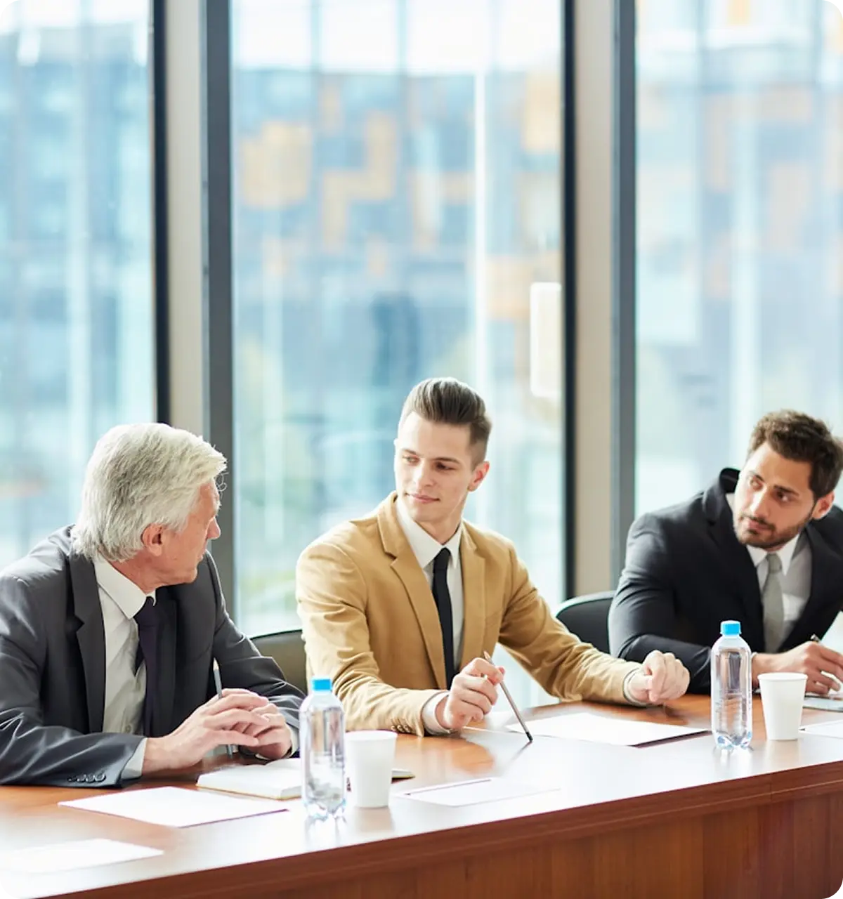 Three men in suits seated at a conference table with water bottles and papers, engaged in a discussion.