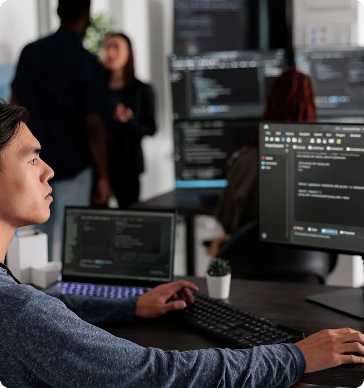 Young man focused on coding at a desk with multiple computer screens displaying code, while colleagues converse in the background of a modern office.