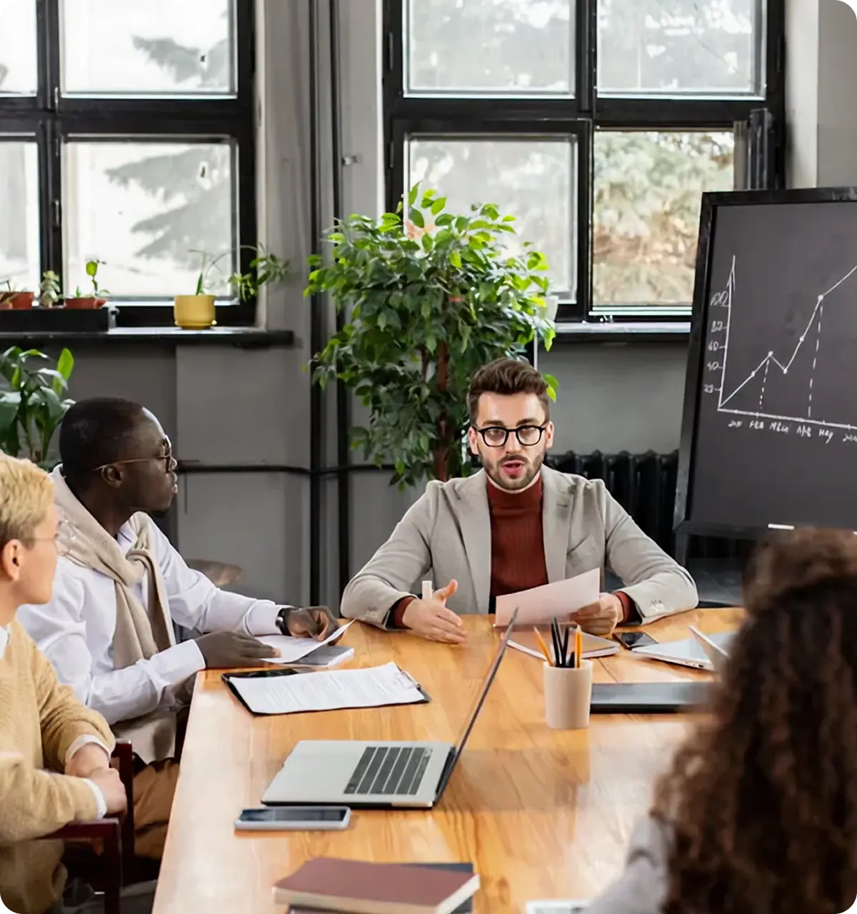 A man in glasses and a blazer speaking while holding a paper in a business meeting with colleagues around a wooden table and a chart on a blackboard.
