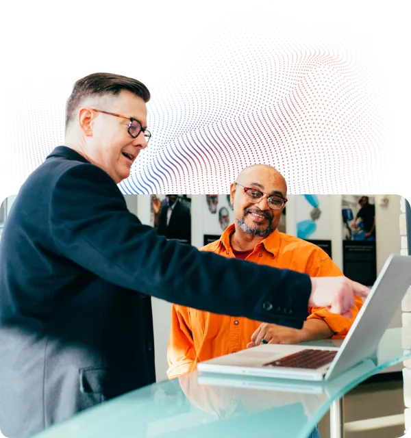 Man in black jacket pointing at a laptop screen while another man in an orange shirt watches and smiles.