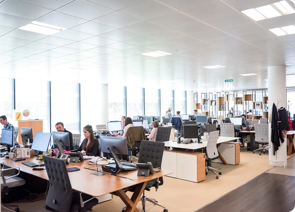 Modern open-plan office with multiple employees working at desks with computers and office supplies.
