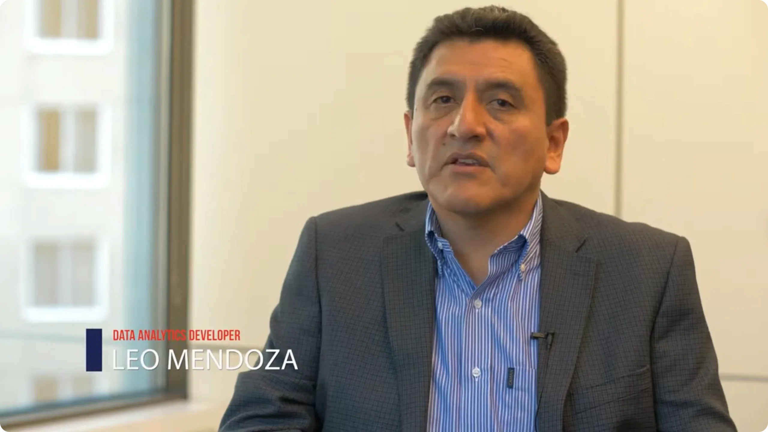 Man in a checkered blazer and blue striped shirt sitting indoors next to a window, identified as Leo Mendoza, Data Analytics Developer.