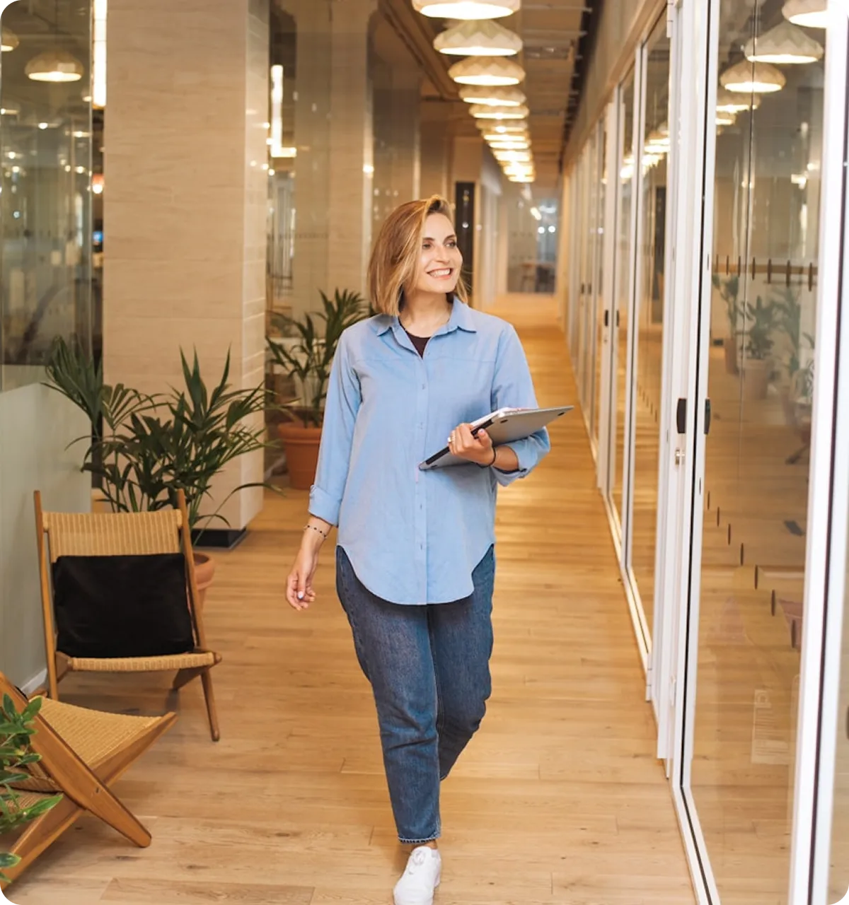 Smiling woman in casual blue shirt holding a laptop walking in a modern office hallway with wooden floors and glass walls.