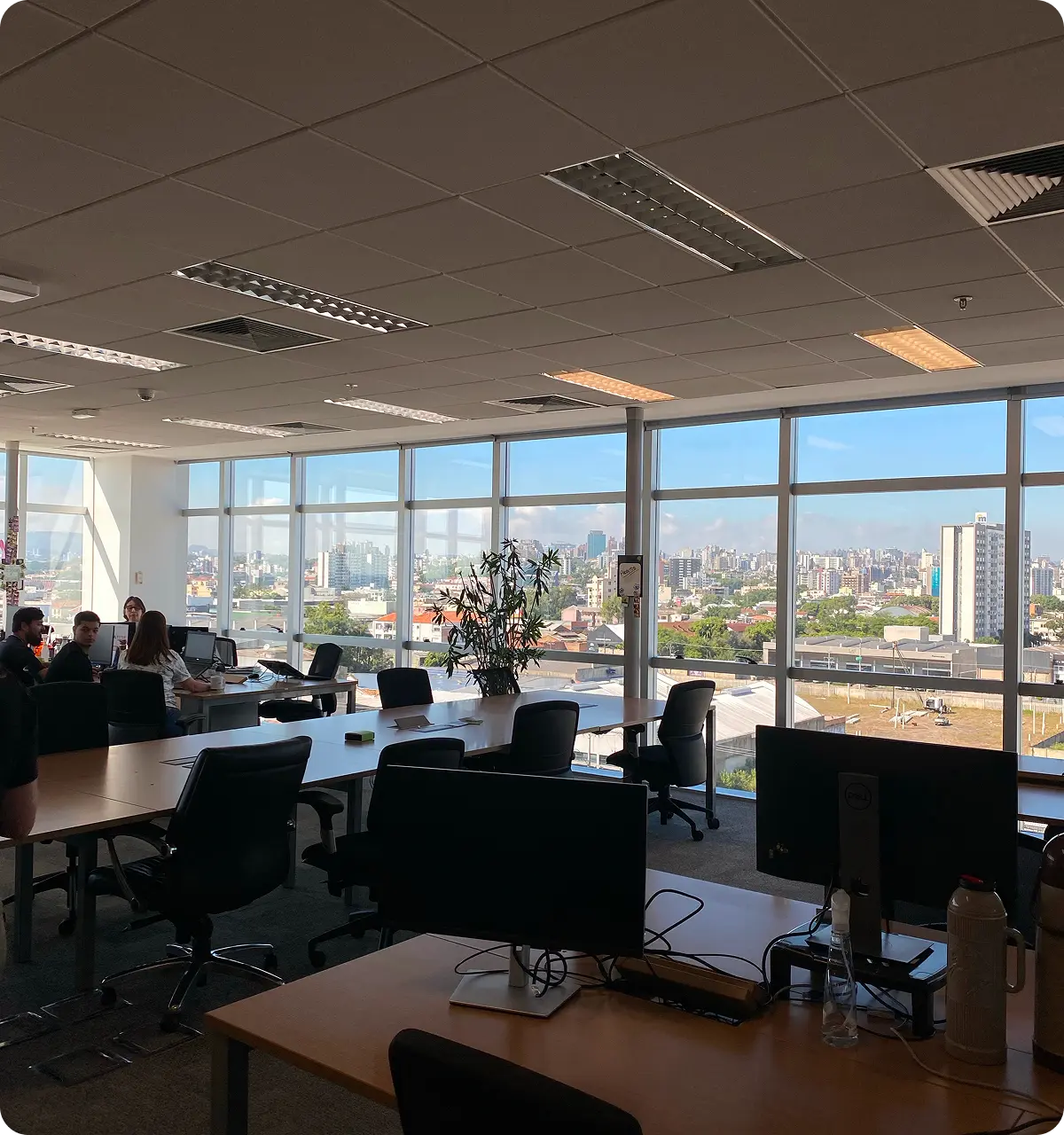 Modern office space with large windows overlooking a city skyline, several desks and chairs, and a few people working.
