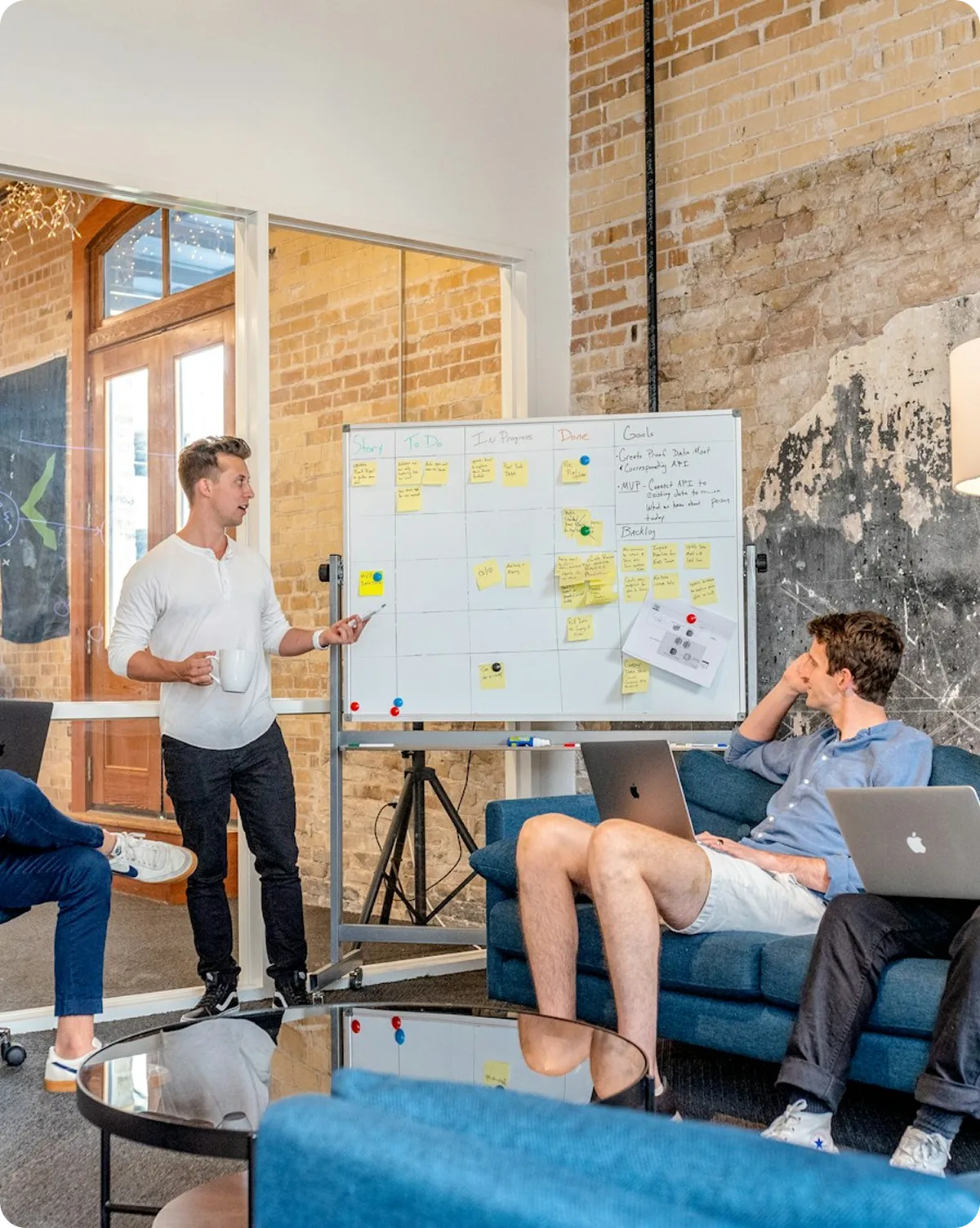 Man standing and presenting with a marker in front of a whiteboard covered in sticky notes while two seated men with laptops listen.