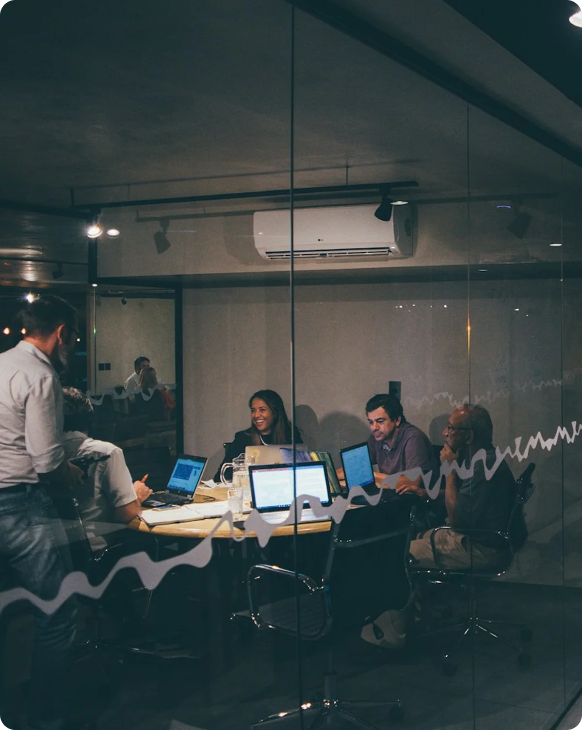 Five people having a meeting around a table with laptops in a glass-walled conference room.