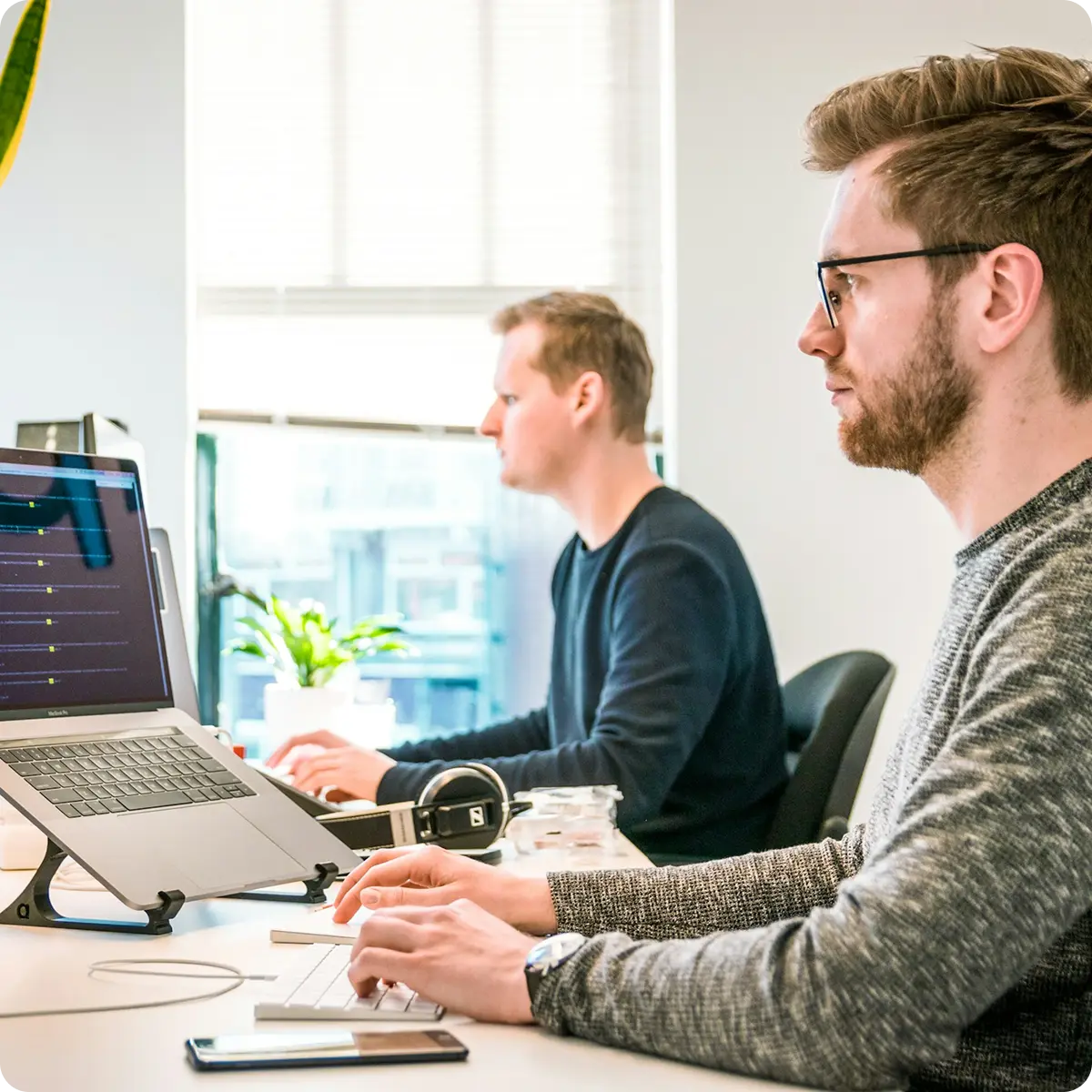 Two men working on laptops at a desk in a bright office with a window and a plant in the background.
