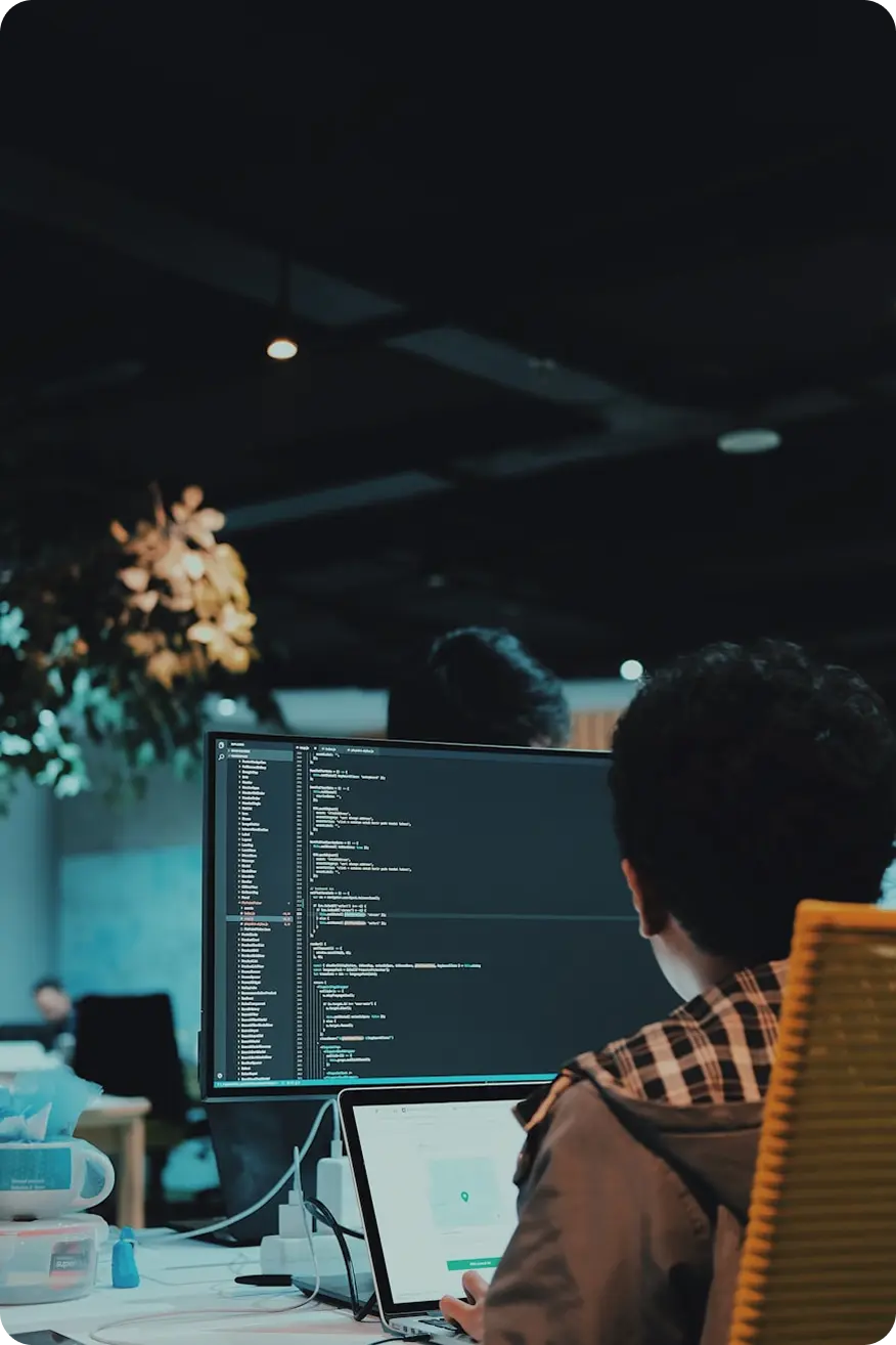 Person working on a laptop with a large monitor displaying programming code in a dimly lit office.