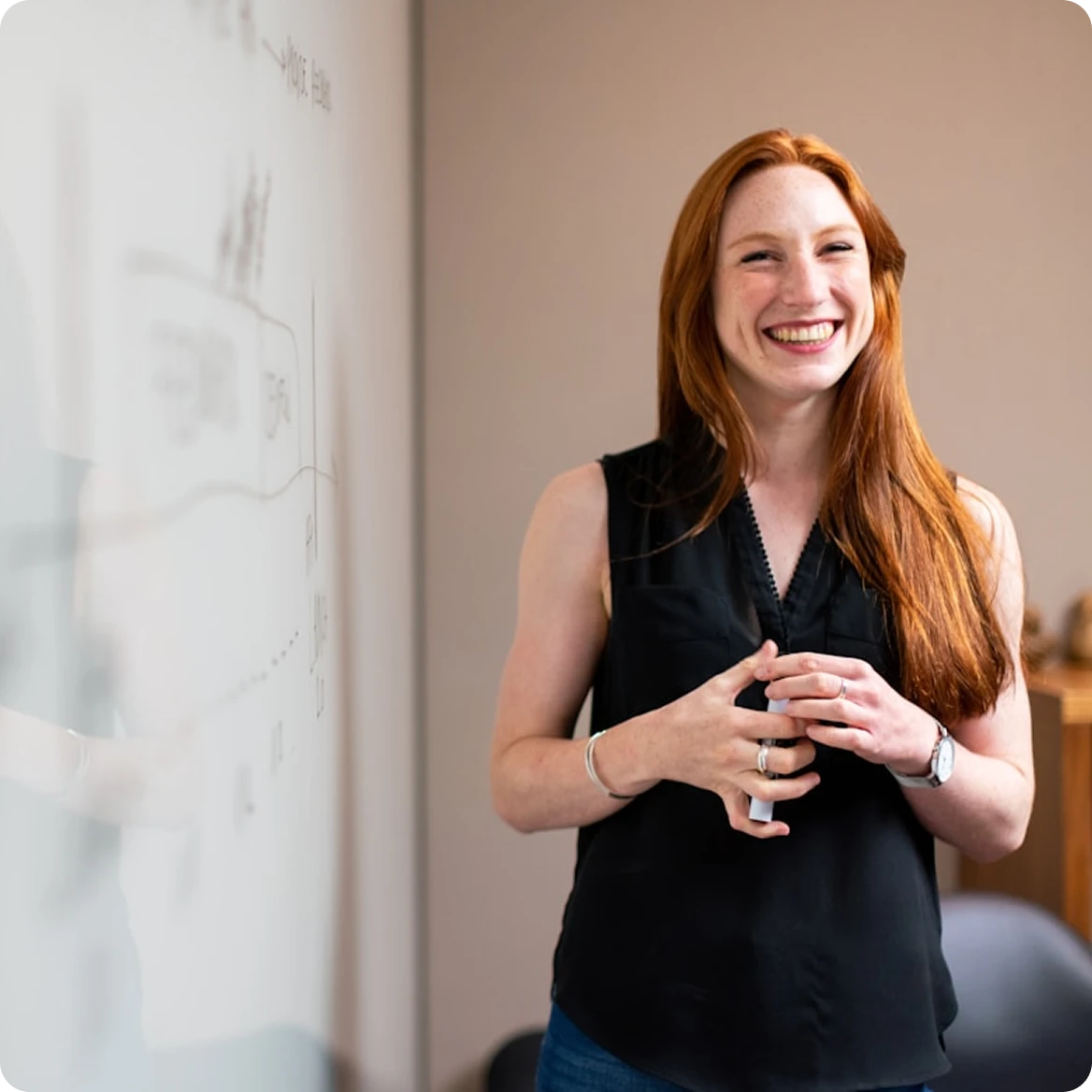 Smiling woman with long red hair standing near a whiteboard in an office setting.
