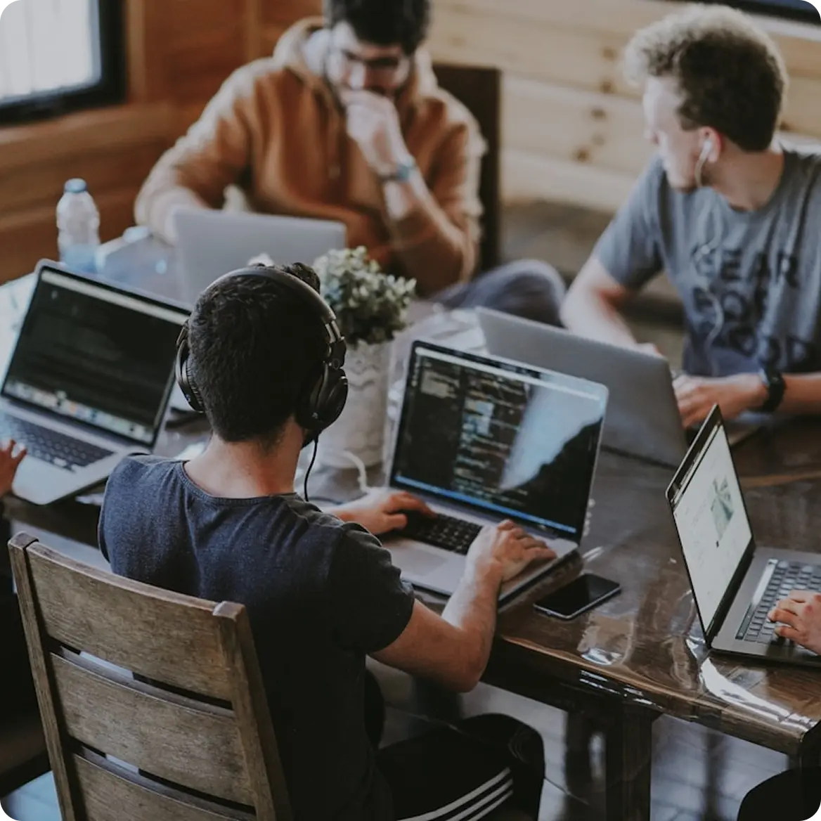 Four people working on laptops around a wooden table in a cozy room, with one person wearing headphones.