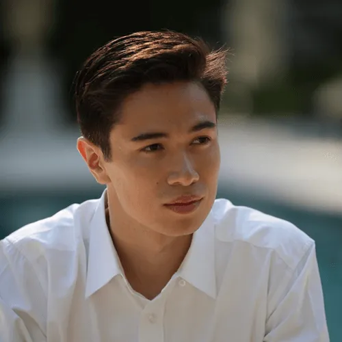 Young man with dark hair wearing a white shirt, looking contemplatively to the side against a blurred background.