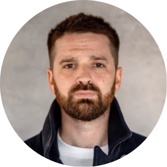 Man with short brown hair and beard wearing a dark jacket over a white shirt against a gray background.