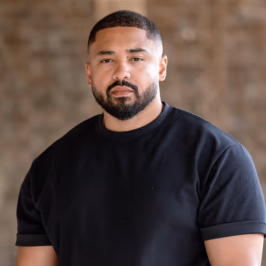 Portrait of a man with short hair and beard wearing a black rolled-up sleeve t-shirt against a blurred brown background.